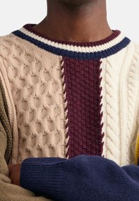 Uomo che indossa un maglione irlandese multicolore a trama cable-knit con sezioni beige, bordeaux, crema, oliva e blu navy, con le braccia incrociate.