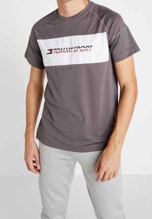 Hombre vistiendo una camiseta de manga corta Tommy Sport gris con una franja horizontal blanca, combinada con pantalones deportivos gris claro sobre un fondo liso.