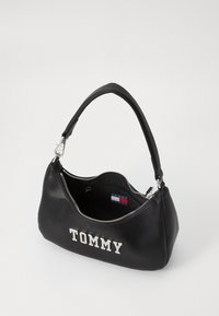 Svart syntetisk håndveske med en buet silhuett, sølvglitrende glidelås og preget "TOMMY"-logo på forsiden, med en teksturert stropp og innvendig lomme.
