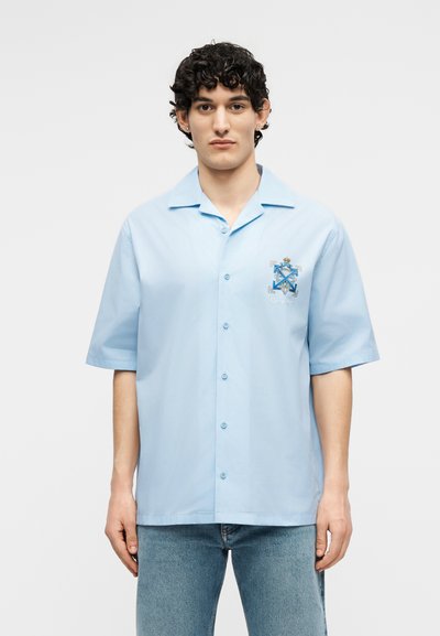 Νεαρός άνδρας με ανοιχτό μπλε πουκάμισο με κοντά μανίκια, γιακά και κεντημένο έμβλημα στο αριστερό στήθος, συνδυασμένο με μπλε τζιν.