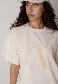 Jasnobeżowa koszulka wykonana z miękkiej tkaniny, z dużym jasnobeżowym logo "A" w kole, z krótkimi rękawami i okrągłym dekoltem.