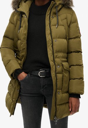 Osoba ubrana w oliwkowozieloną pikowaną parkę z kapturem wykończonym futerkiem, nałożoną na czarny sweter i ciemne dżinsy, z ręką w kieszeni.
