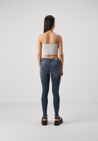 Kvinna med långt mörkt hår, iförd en grå kortlinne, smala blå jeans och svarta plattformsandaletter, stående mot en enkel bakgrund.