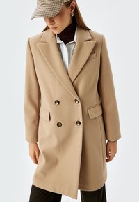 Abrigo beige de doble botonadura con solapas de pico, dos bolsillos delanteros y botones oscuros. Textura suave, corte recto, combinado con un suéter de cuello alto marrón.