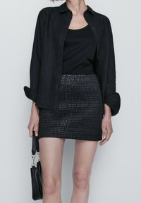 Zwarte linnen overhemd met knopen over een zwarte tanktop, gecombineerd met een textuur zwart mini-skirt met een gekreukt patroon. Inclusief zwarte handtas.