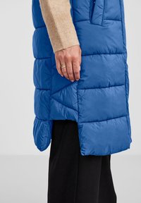 Mėlynas pūkinis liemenė su siuvinėtu raštu, šoniniu įpjovimu ir lygiu paviršiumi. Ant liemenės šoninio siūlo guli ranka su auksiniu žiedo.