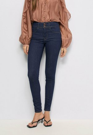 Mujer con vaqueros ajustados de talle alto en azul oscuro, una blusa marrón brillante de manga larga y sandalias negras de tacón con tiras finas.