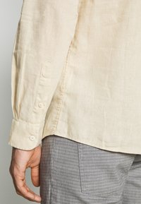 Bras d'homme portant une chemise beige à manches longues avec poignet boutonné et un pantalon gris texturé, debout devant un fond neutre.