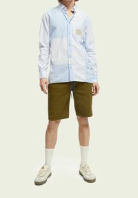 Camicia a righe azzurre e bianche con bottoni e taschino; abbinata a pantaloni corte verde oliva e calzini chiari, completati da sneaker neutre.