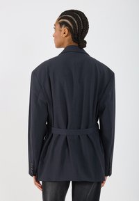Blazer de cărbune cu un design structurat, având umeri supradimensionați, talie cu curea și manșete cu nasturi. Material din lână netedă cu o textură subtilă.