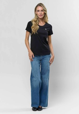 Čierne bavlnené tričko s okrúhlym výstrihom, nosené s svetlomodrými rozšírenými džínami. Má malé logo na hrudi a minimálne prsty v švy.