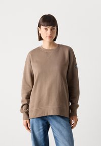 Brun genser med rund hals og lange ermer, med ribbestrikkede mansjetter og et sidemerke. Båret med lyseblå jeans.