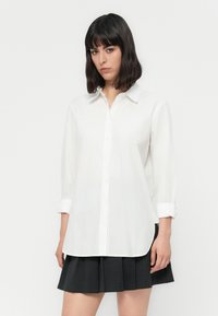 Camisa branca de manga longa com colarinho clássico, com as mangas arroladas nos punhos e uma bainha arredondada. Tecido suave com um corte relaxado.