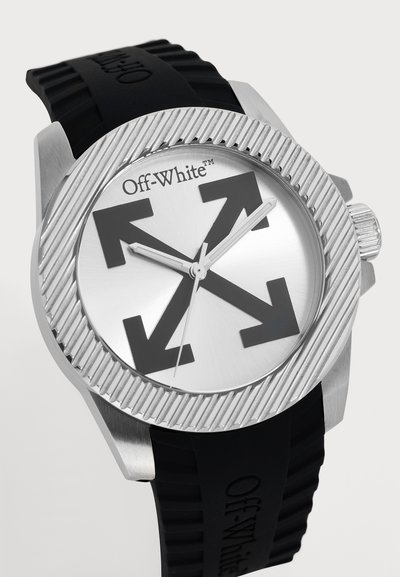 Gros plan sur une montre-bracelet argentée Off-White, avec une lunette cannelée, un motif de flèche marqué sur le cadran et un bracelet en caoutchouc noir.