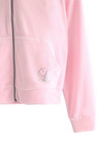 Jasnoróżowa bluza na zamek z miękkiego materiału z dwiema przednimi kieszeniami. Posiada srebrny akcent w postaci monogramu z dżetów po lewej stronie.