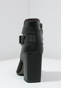 Botín negro de cuero hasta el tobillo con un alto tacón cuadrado, que presenta una textura a rayas, una correa ancha y un detalle de hebilla en el costado.