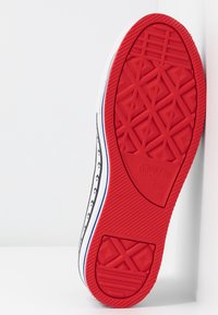 Suola rossa in gomma di una sneaker bianca Converse All Star con motivo a stelle nere sul lato, inclinata contro uno sfondo bianco.