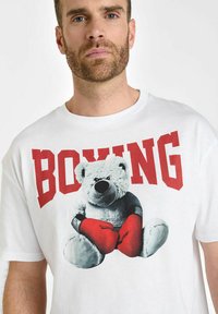 Camiseta blanca con un gráfico de un oso en guantes de boxeo rojos, con la palabra "BOXING" en letras negritas rojas encima del oso.