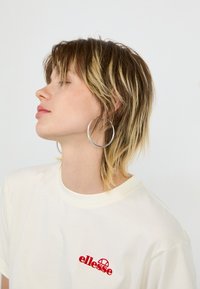 T-shirt en coton blanc avec un petit logo "ellesse" rouge, porté avec de grandes boucles d'oreilles en argent. Les cheveux sont ébouriffés, montrant une coupe en couches.