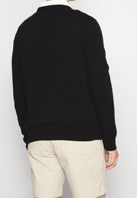 Man i svart långärmad tröja och beige shorts, stående vänd bort från kameran mot en enfärgad vit bakgrund.