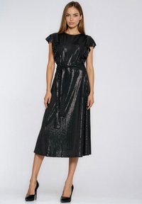 Kobieta ubrana w błyszczącą czarną sukienkę midi z falbankowanymi rękawami i wiązaniem w talii, w połączeniu z czarnymi wysokimi obcasami, stojąca na jednolitym tle.
