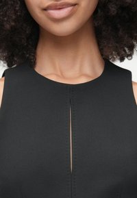 Čierne bezrukávové šaty s okrúhlym výstrihom a vertikálnym rozparkom na hrudi, s jemnými šitými detailmi na pleciach.