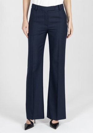 Kobieta ubrana w granatowe, szerokie, dopasowane spodnie i czarne szpilki z ostrym noskiem, stojąca na jednolitym białym tle.