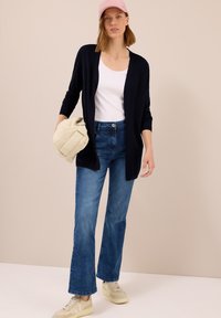 Marineblauer Cardigan über einem weißen Oberteil, blaue Jeans und beige Sneakers. Eine hellrosa Kappe wird gehalten. Einfaches Design ohne sichtbare Muster.
