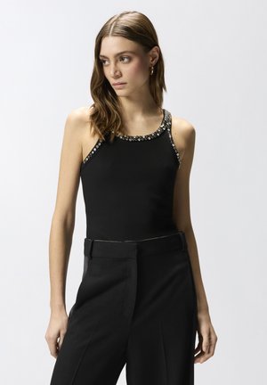 Γυναίκα με ανοιχτό καστανά μαλλιά φοράει μαύρο αμάνικο τοπ με ασημένια καρφιά στο λαιμό και μαύρο ψηλόμεσο παντελόνι, κοιτάζοντας πλάι.