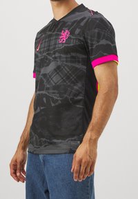 Maglietta nera a maniche corte con un motivo grafico testurizzato, accenti rosa sulle maniche e uno stemma con un leone sul petto.