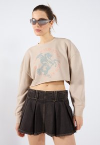 Felpa beige a maniche lunghe con grafica di un cavallo blu e arancione, abbinata a una minigonna plissettata in denim nera con orlo sfilacciato.