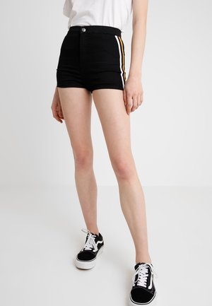 Shorts de mezclilla negra de tiro alto, con franjas laterales en amarillo y blanco. Combinados con zapatillas negras y blancas.