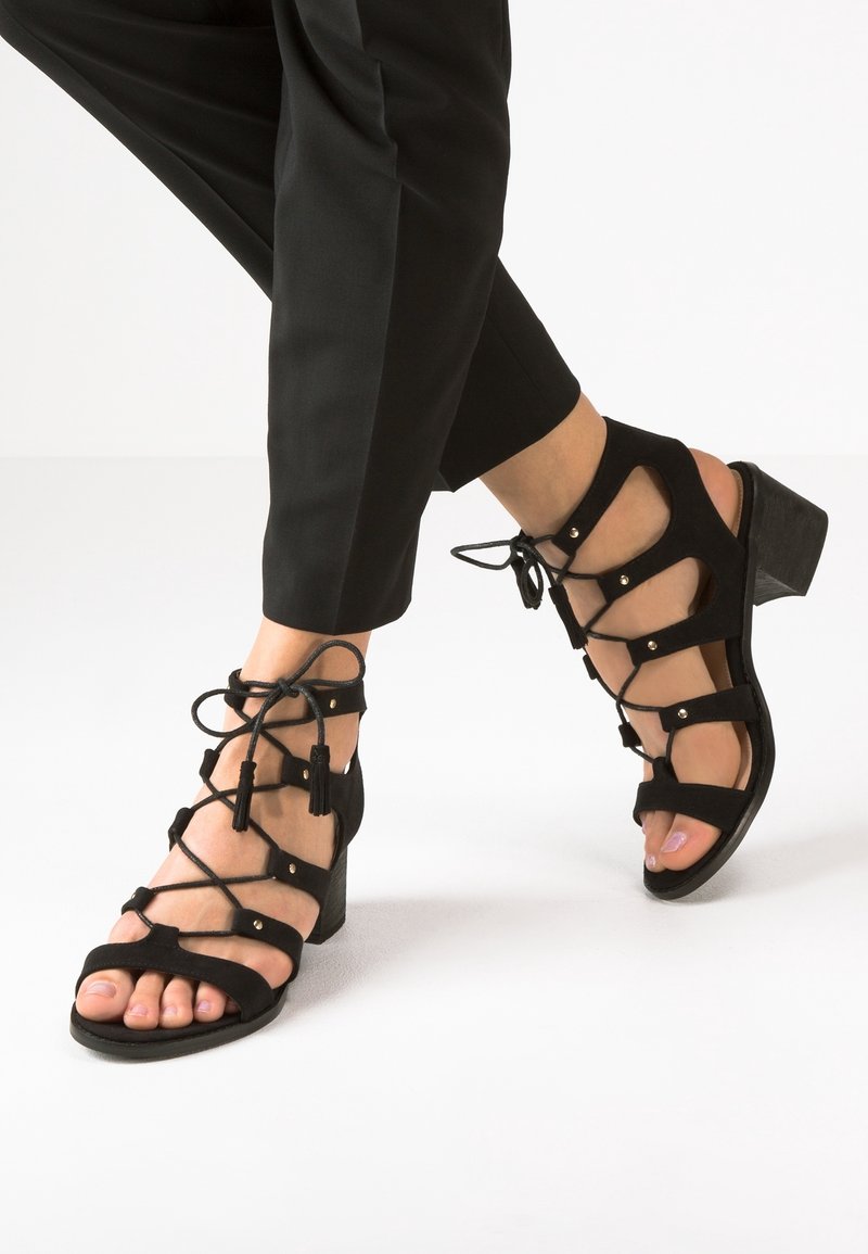 Sandales à talons en daim noir avec des brides entrelacées, rehaussées de petits clous dorés et de glands, dotées d'un bout ouvert et d'un talon épais.