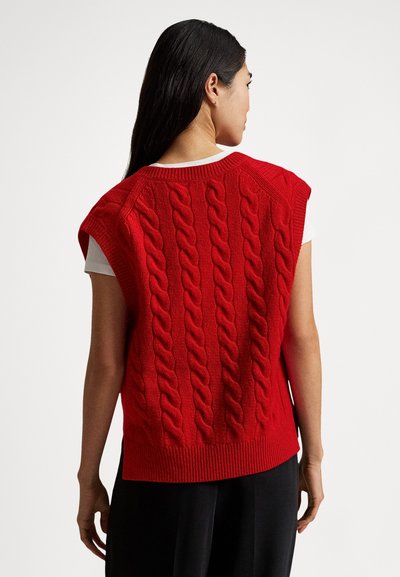 Rdeč pleten brezrokavnik z vzorcem kitke, rebrastim izrezom in robom. Nošen čez belo srajco. Gladek videz, sproščen kroj.