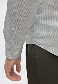 Gros plan sur le bras gauche d’une personne portant une chemise rayée à manches longues avec des boutons blancs et un pantalon sombre.