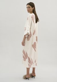 Robe longue et fluide en crème avec un motif de coups de pinceau abstraits roses. Tissu doux, manches larges et fentes sur les côtés. Portée avec des talons accentués de fourrure.