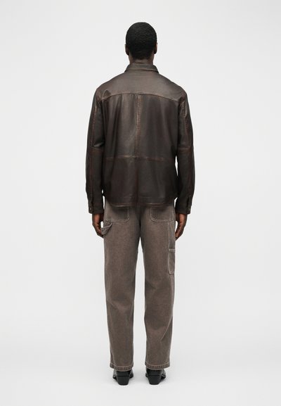 Άνδρας που φοράει σκούρο καφέ δερμάτινο μπουφάν και γκρι παντελόνι cargo, στέκεται με την πλάτη προς τα εμπρός σε απλό λευκό φόντο.