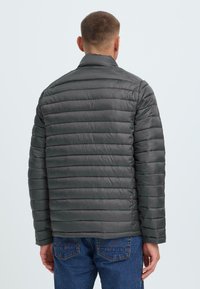 Grå polstret jakke med horisontalt quiltet design og stående krage. Har lange ermer og en jevn, blank tekstur.