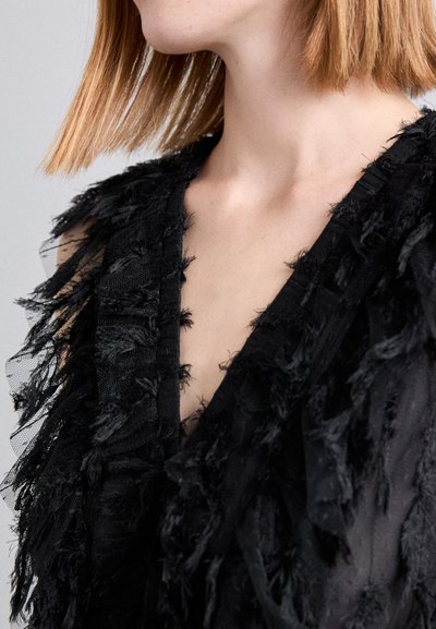 Vêtement noir avec un décolleté en V profond, présentant des franges texturées le long des bords et un design en couches. Fabriqué en tissu transparent avec une finition douce.