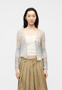 Γυναίκα που φοράει ένα παστέλ ριγέ πλεκτό ζακέτα πάνω από λευκό πλεκτό κοντό μπλουζάκι και μπεζ πλισέ φούστα, στέκεται μπροστά σε απλό φόντο.