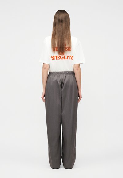 Άτομο με μακριά ίσια μαλλιά, γυρισμένο προς τα πίσω, φοράει λευκό μπλουζάκι με πορτοκαλί κείμενο και φαρδιά γκρι παντελόνια.