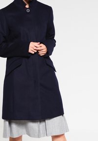 Marinblå ullrock med hög krage, knäppning fram och sidofickor. Har en skräddarsydd passform och slät textur, bärs över en grå plisserad kjol.