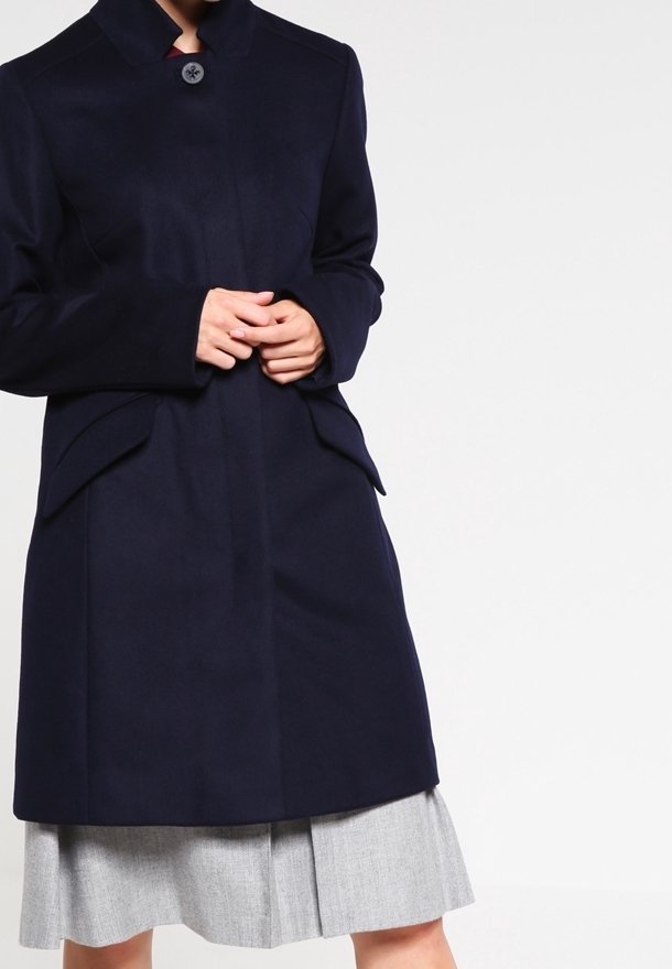Marinblå ullrock med hög krage, knäppning fram och sidofickor. Har en skräddarsydd passform och slät textur, bärs över en grå plisserad kjol.