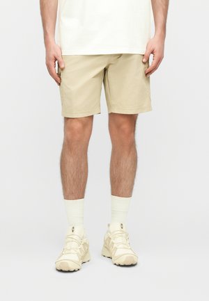 Mees, kes kannab beežikaid põlveni ulatuvaid lühikesi pükse, valgeid kummisukke ja kreemjat värvi spordijalatseid, seisab ühevärvilise tausta ees.