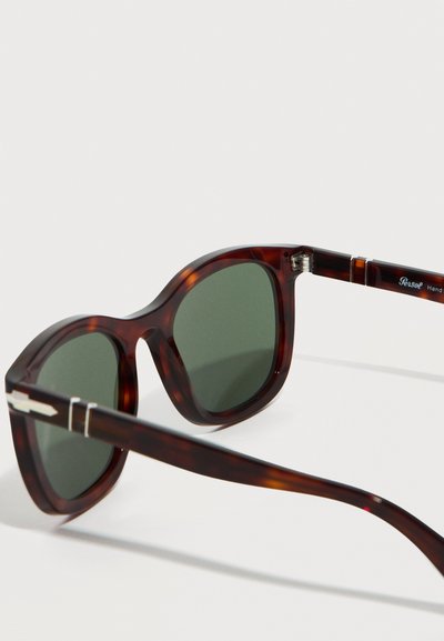 Γυαλιά ηλίου με κορνίζα σε χρώμα χελώνας, φακούς σκούρου πράσινου και ασημένιες λεπτομέρειες στα κλαδιά, ακουμπισμένα σε λευκή επιφάνεια.