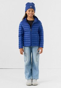 Blauwe donsjas met ritssluiting aan de voorkant en capuchon, gecombineerd met een zwart shirt en lichtblauwe cargobroek. Het model draagt een blauwe muts en grijze pantoffels.