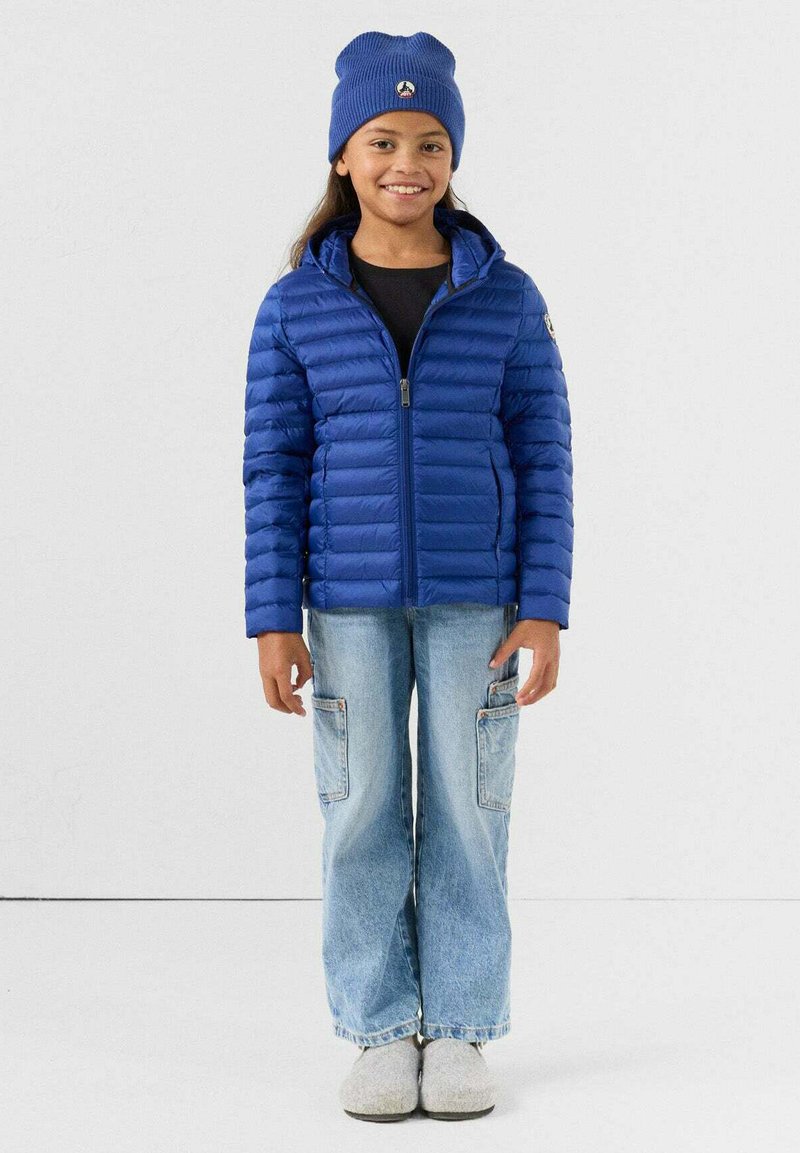 Blauwe donsjas met ritssluiting aan de voorkant en capuchon, gecombineerd met een zwart shirt en lichtblauwe cargobroek. Het model draagt een blauwe muts en grijze pantoffels.