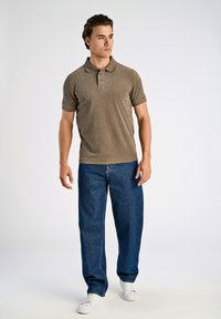Polo a maniche corte da uomo in tessuto beige, abbinato a jeans blu larghi. Sneakers bianche completano il look casual.