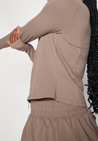 Long-sleeve, light brown top made of smooth fabric with a loose fit. Features a rounded hem and is paired with a matching elastic-waist skirt.