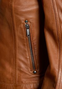 Veste en cuir marron avec un gros plan sur une poche zippée, fermeture éclair en métal argenté et détail en bouton pression noir. Surface douce et texturée.